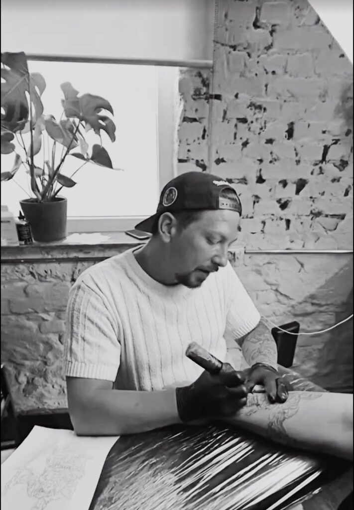 Mann mit neutralem Blick in weißem Shirt tätowiert Arm in grau schwarzem Studio