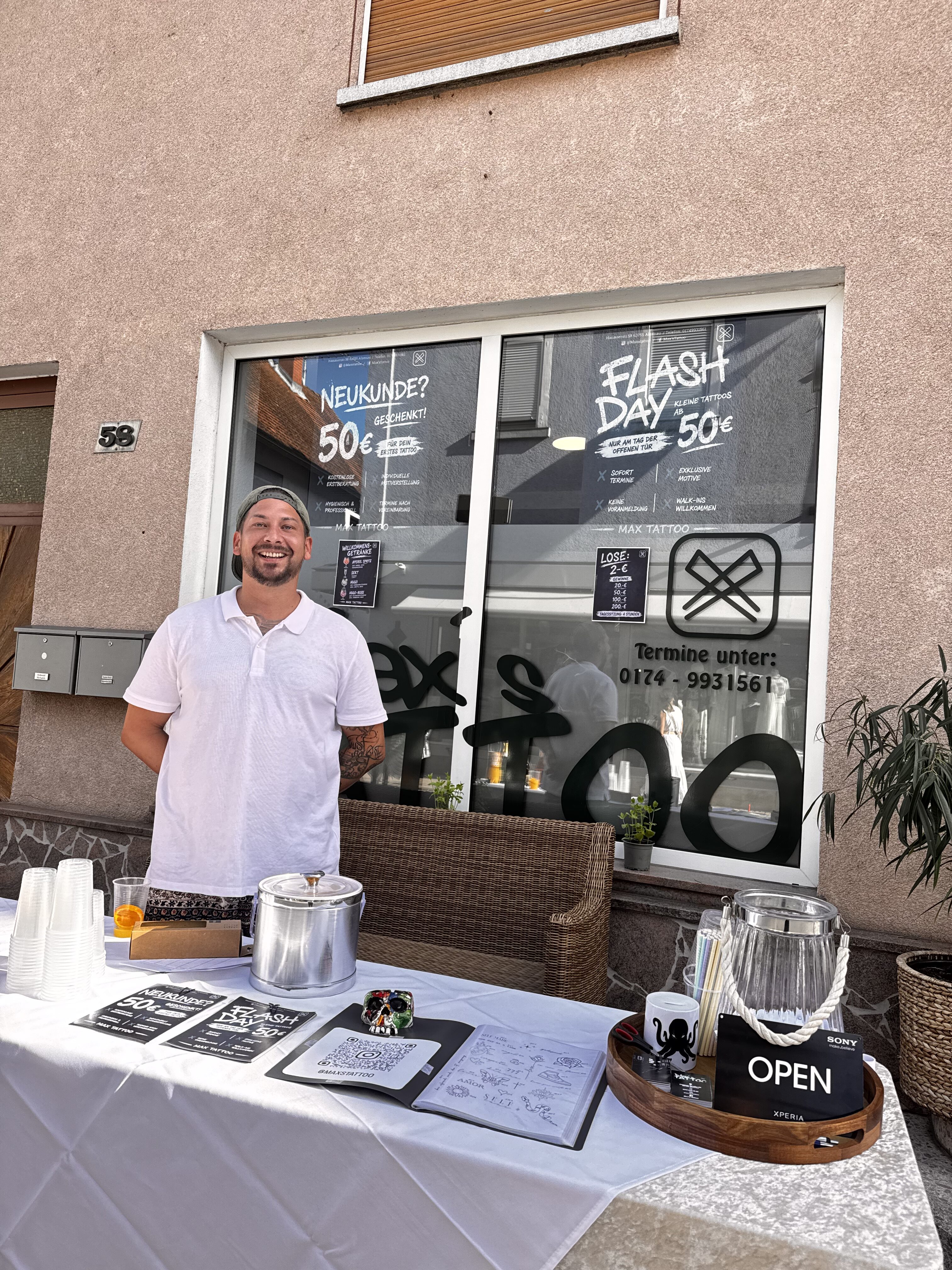 Laechelnder Mann mit Cap und weissem Shirt vor Tattoostudio am Infostand
