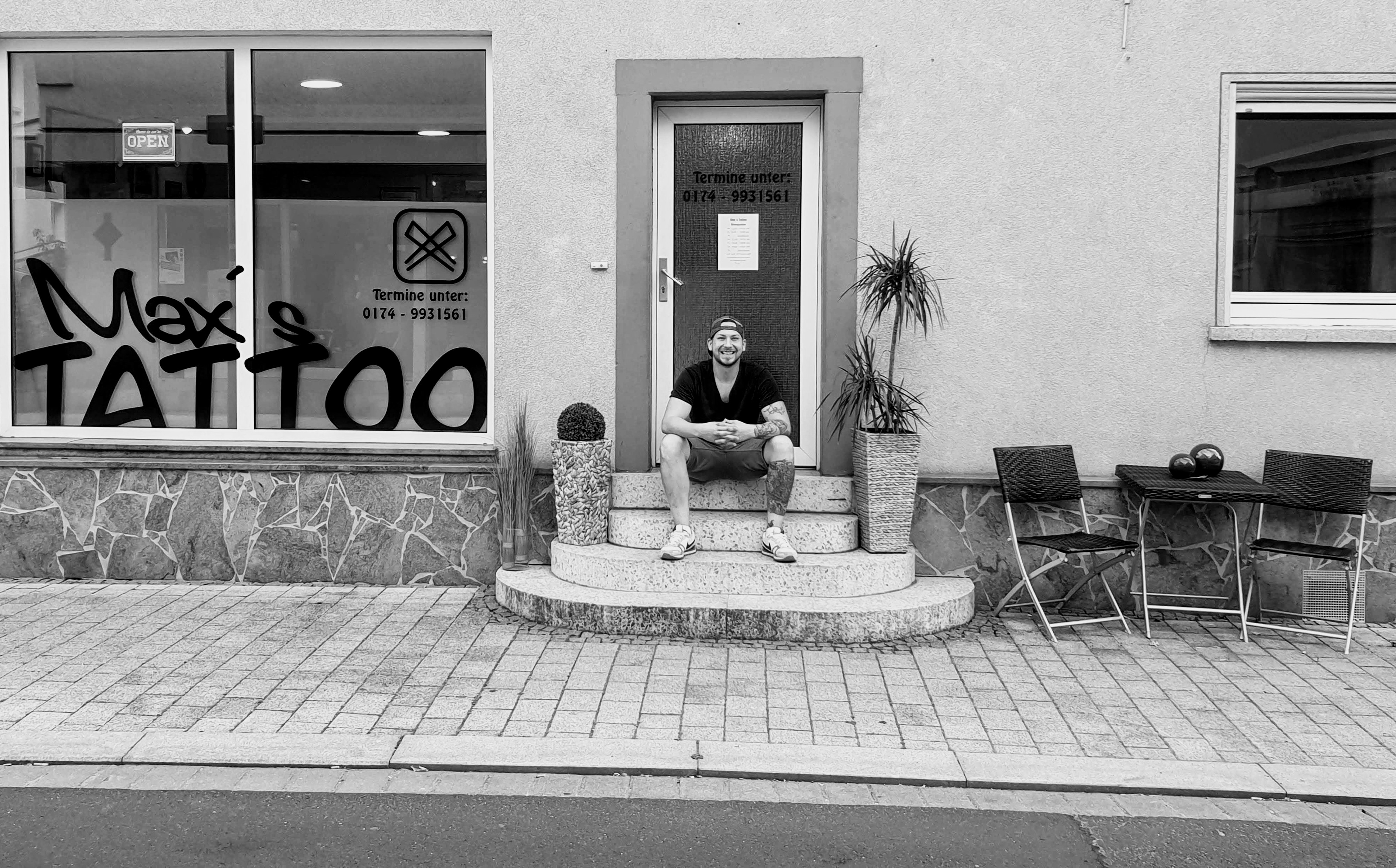 Laechelnder taetowierter Mann mit Cap sitzt vor Max Tattoo Studio