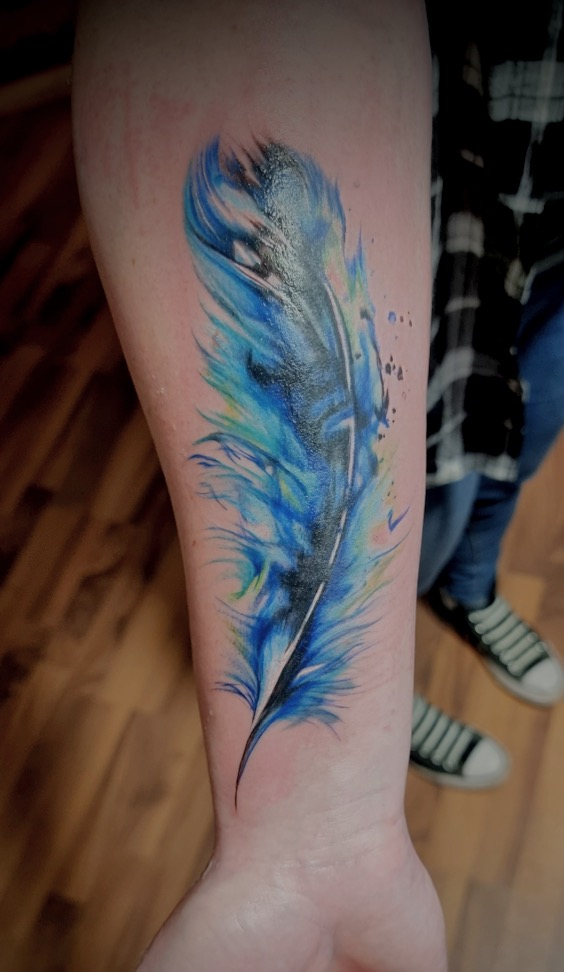 Blaues Feder Tattoo im Aquarellstil auf einem Unterarm mit Farbspritzern